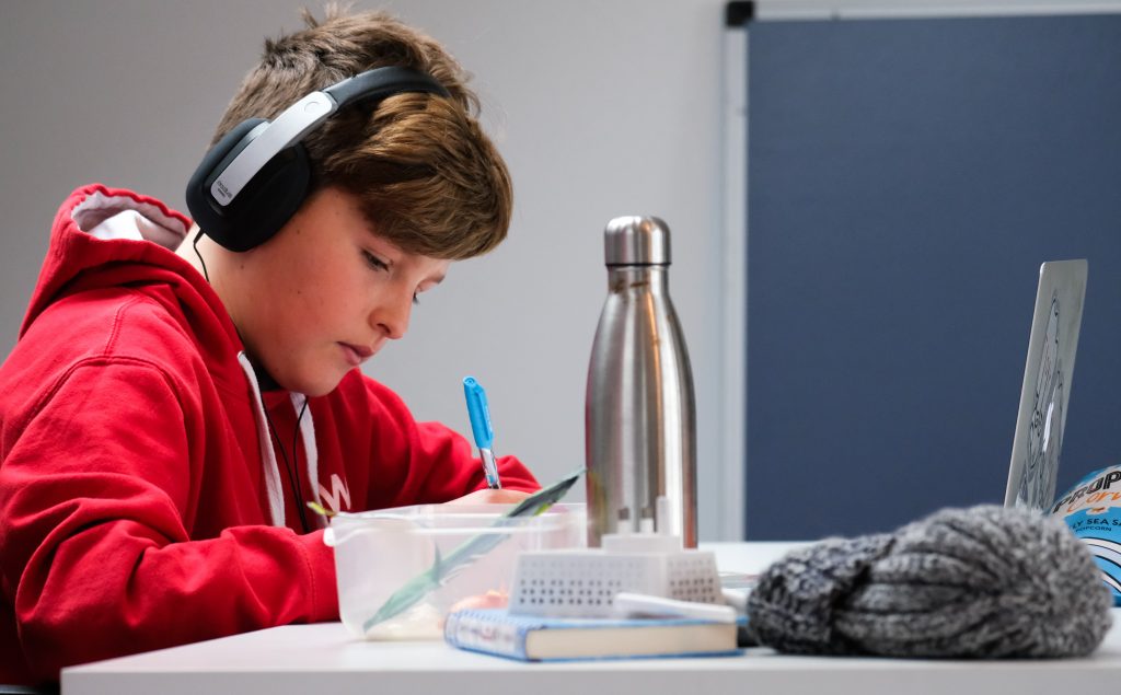 A focused young student wearing a red hoodie and headphones is engaged in studying, taking notes from a laptop screen in a modern classroom setting. Personal items like a water bottle and a knit hat suggest a comfortable and personalized learning environment.