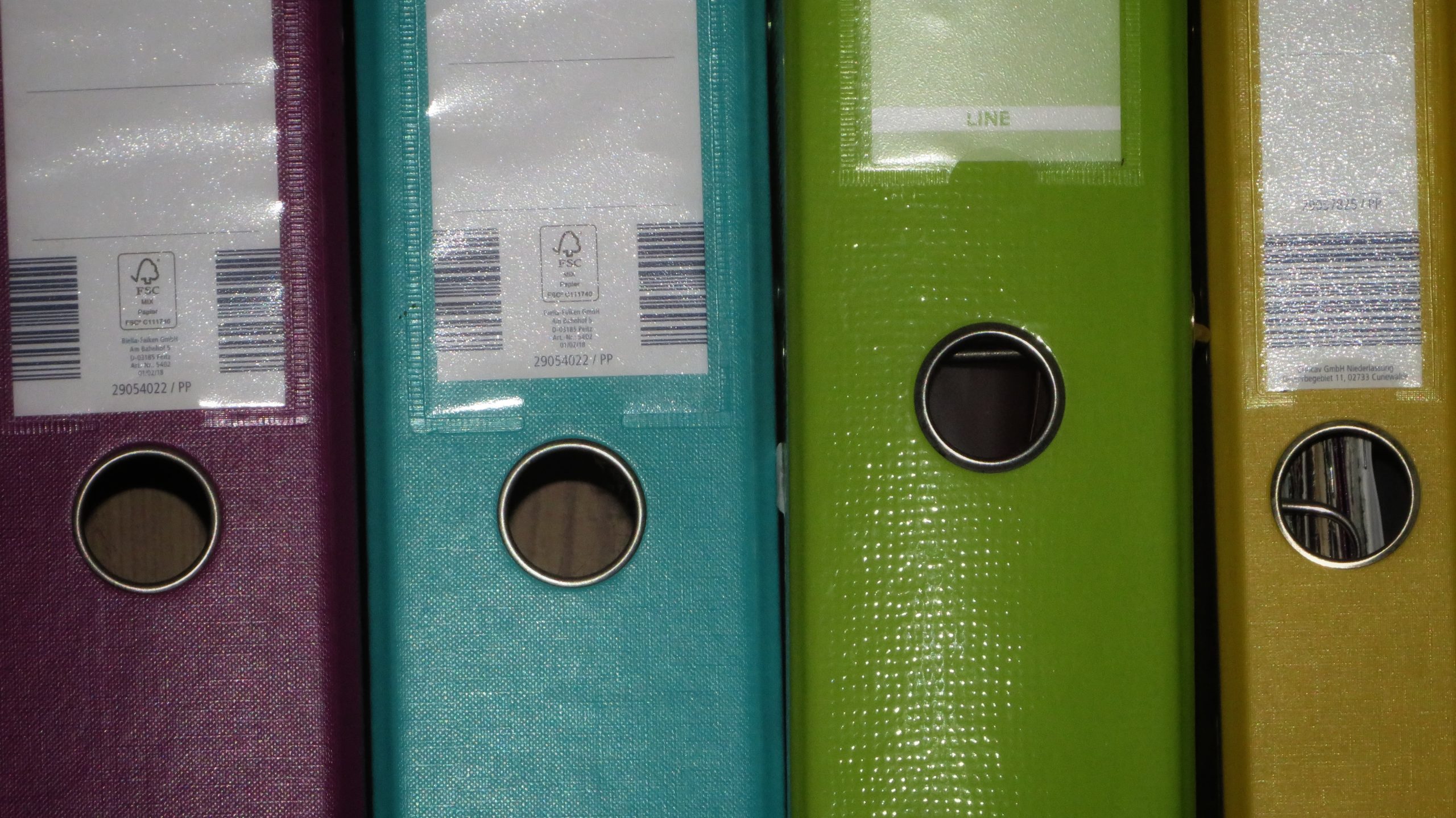 The image shows a close-up of four upright lever arch files in different colors—pink, teal, lime green, and yellow. Each binder has a metal ring at the bottom of the spine for easy handling and a label slot at the top. The textured surfaces of the binders reflect light, and barcode labels are visible on the front covers.