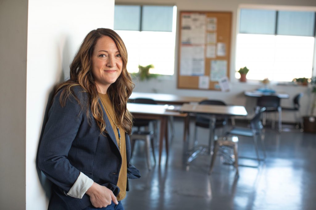 A confident woman with a warm smile leans against a wall in a sunlit classroom, her presence suggesting a supportive and welcoming educational environment. The background, softly out of focus, features tidy desks and informative displays, hinting at a space where learning and growth are fostered.