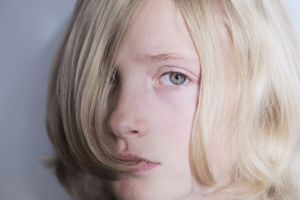 The image shows a close-up of a child with blonde hair partially covering their face, revealing one eye. The child's gaze is directed towards the camera, and the background is a plain, light color, putting the focus on the child's expression.