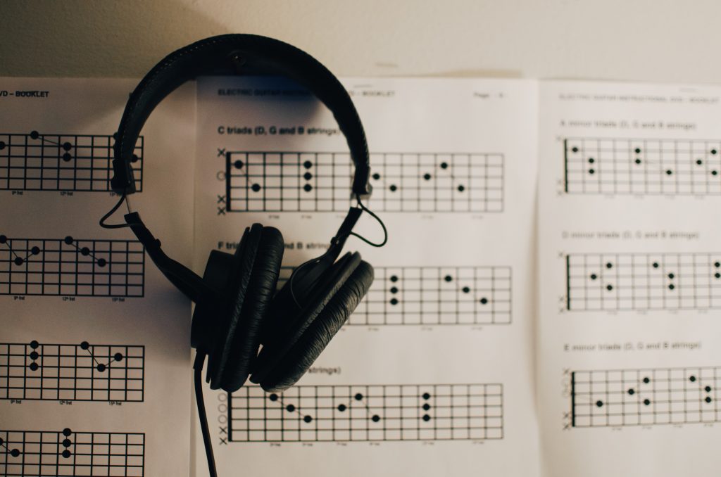 A pair of headphones hangs in front of a wall adorned with sheets of music for electric guitar instruction, showcasing chord diagrams for various triads on different strings, suggesting a learning environment focused on musical skill development.