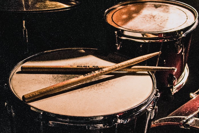 A pair of wooden drumsticks rest on the surface of a snare drum, part of a drum set that includes toms and cymbals, all under a warm, ambient light that highlights the textures and wear of the instruments, suggesting a setting filled with rhythmic energy and musical passion.