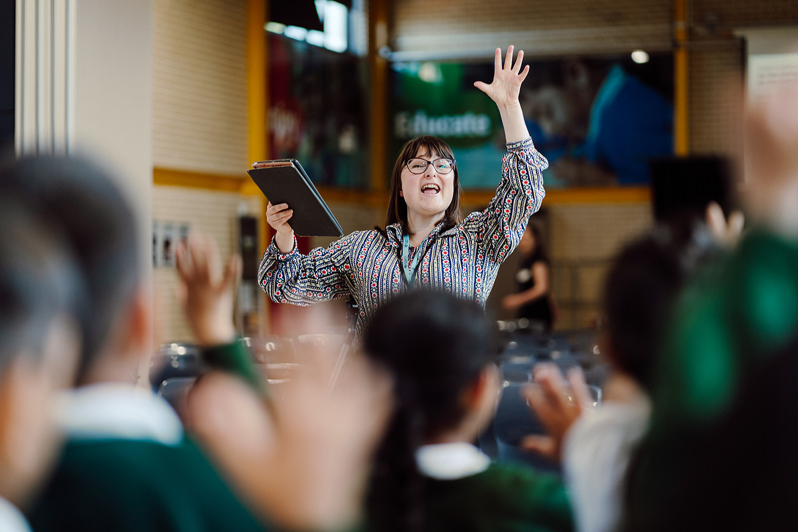 A joyful teacher with glasses and a patterned shirt enthusiastically raises her hand while holding a tablet, leading a classroom of students who are out of focus in the foreground. The setting suggests an interactive and lively educational environment, likely involving music or active participation, as indicated by the students' raised hands responding to the teacher's gesture.