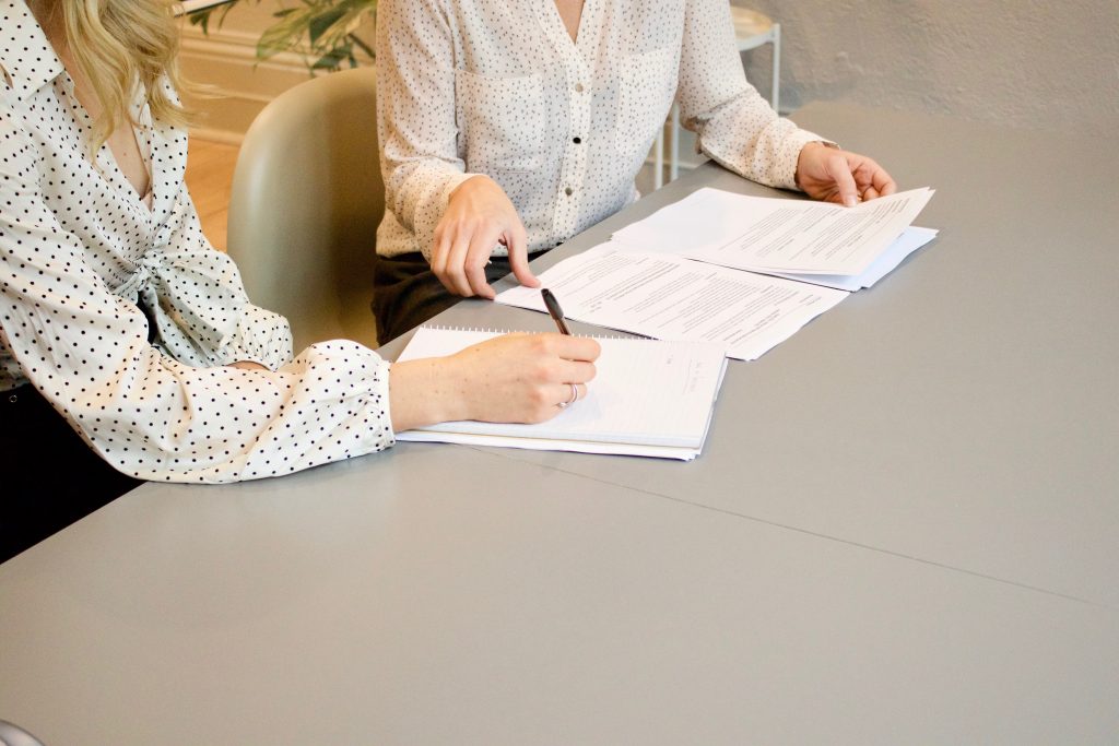 Two individuals are seated at a table, engaged in a review or discussion over documents. Only their torsos and arms are visible. One person is holding a pen and appears to be taking notes on a notepad, while the other is handling some papers. They both wear polka-dotted blouses, suggesting a coordinated or professional setting. The focus is on their collaborative work, with a neutral and calm office environment as the backdrop.