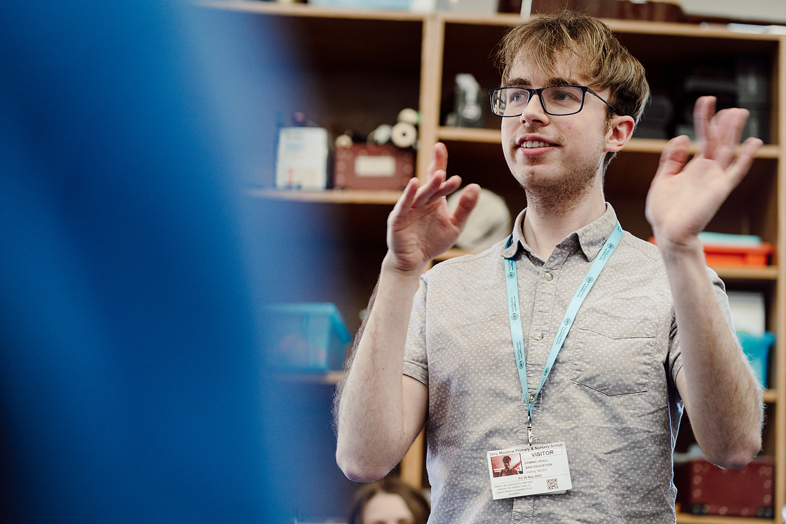 A young male educator with glasses is captured mid-gesture, likely leading a classroom activity or discussion. He's wearing a visitor badge, indicating he might be a guest teacher or specialist. The blurred foreground suggests a student's perspective, adding a sense of engagement and focus on the educator. The classroom setting is indicated by shelves with various items in the background, creating an educational atmosphere.