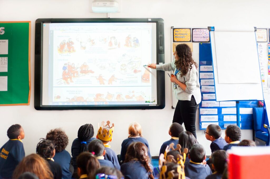 A teacher stands beside an interactive whiteboard, pointing to educational content, while a group of young, attentive students, some wearing paper crowns, sit on the floor, looking up and engaging with the lesson. The classroom environment is bright and colorful, with educational materials visible, fostering a dynamic learning experience.
