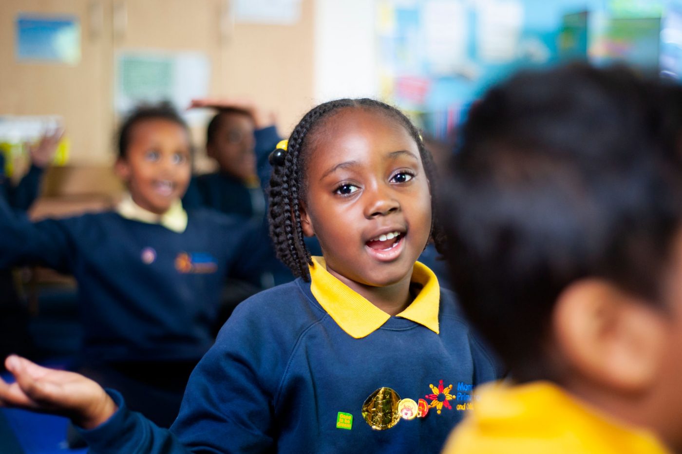 A young girl with braided hair and a bright expression participates enthusiastically in a classroom activity, wearing a navy and yellow school uniform adorned with colorful badges. Her peers, slightly out of focus, share in the lively educational experience, reflecting a vibrant and interactive learning environment.