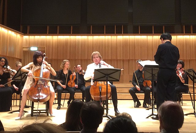 The image shows a group of classical musicians performing on stage. In the foreground, a cellist is prominently seated, playing her instrument, with sheet music on a stand in front of her. Beside her, another cellist is also focused on his performance. In the background, other members of the string section, including violinists, can be seen. The conductor is standing with his back to the camera, leading the ensemble. The audience is visible in the lower part of the image, indicating a live concert setting with a wooden-paneled concert hall enhancing the ambiance.