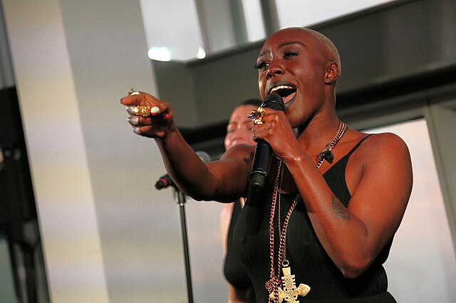 A vocalist passionately performs, her hand extended towards the audience as she sings into a microphone, conveying a moment of musical expression and connection. Behind her, another singer is partially visible, suggesting a shared performance or chorus. The focus on the main singer captures the intensity and emotion of live music.