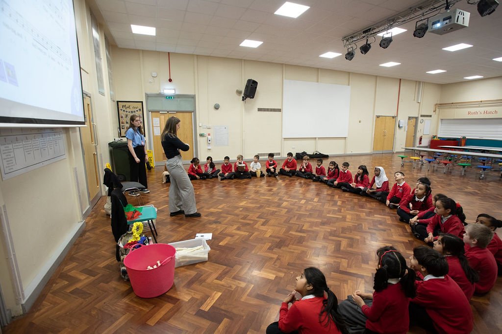 A spacious school hall filled with attentive children seated on the floor forms a semi-circle around two instructors, one of whom is actively engaging with the students. A projector screen displays musical notation, suggesting a lesson in progress, while various musical instruments and colorful props are scattered nearby, indicating an interactive and creative learning environment. The atmosphere is one of focused learning, with the children dressed in uniform, embodying a typical school setting.