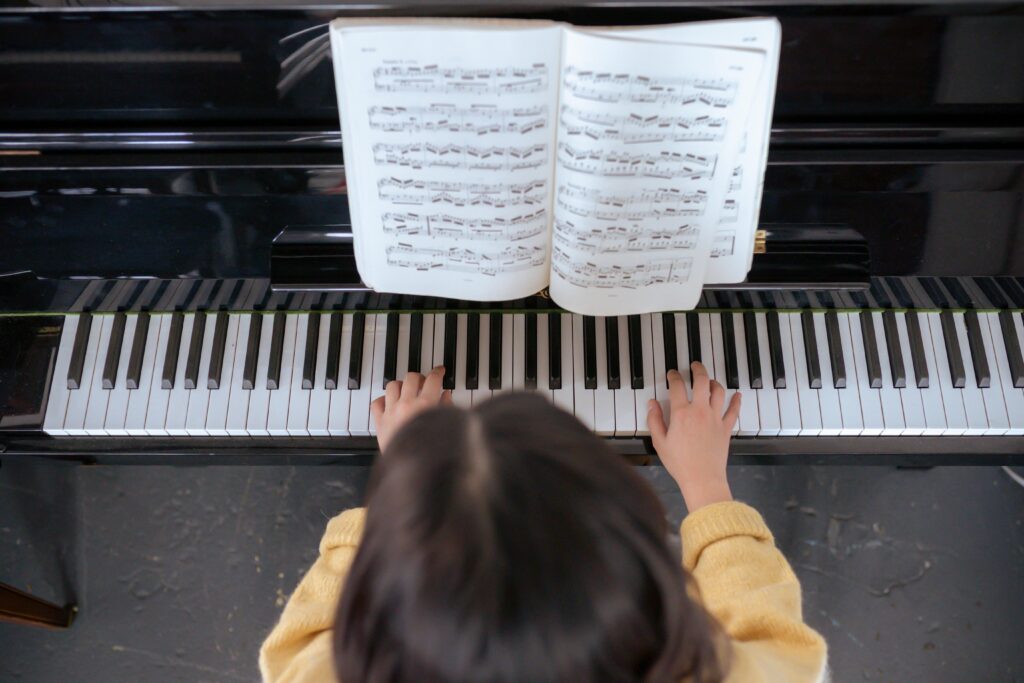 A child in a yellow sweater is practicing piano, with their focus on a sheet music book propped open on the music stand. The perspective is from above, capturing the child's hands on the keys and the reflection of the music notes on the polished surface of the piano.