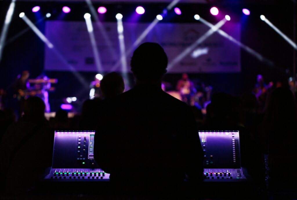 A silhouette of a person is seen from behind, overseeing a live concert from a sound mixing console, with colorful stage lights illuminating musicians performing in the background. The atmosphere suggests a blend of focus and creativity, integral to delivering a high-quality live music experience.