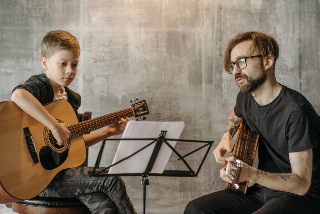 A young child and an adult are seated, each holding an acoustic guitar, focused on their instruments. The child appears to be playing, looking intently at the strings, while the adult watches, ready to guide or play along. A music stand with sheet music is positioned between them, suggesting a learning or practice session. The background is a textured wall, providing a calm and neutral setting for the musical interaction.