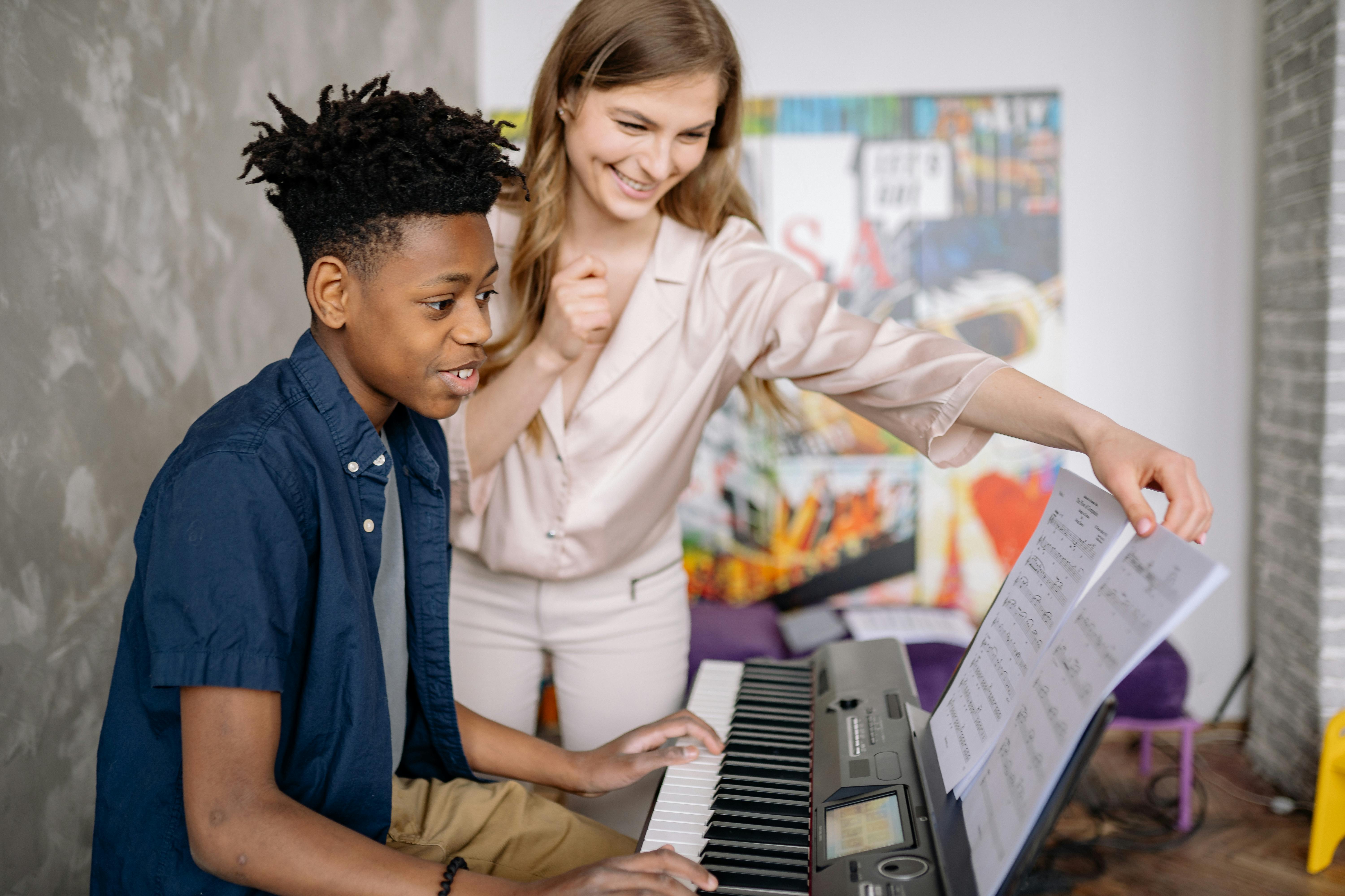 A young student with a focused expression plays a keyboard while a smiling teacher stands beside him, gesturing encouragingly and holding a sheet of music. The background features colorful artwork, suggesting a creative and supportive learning environment.