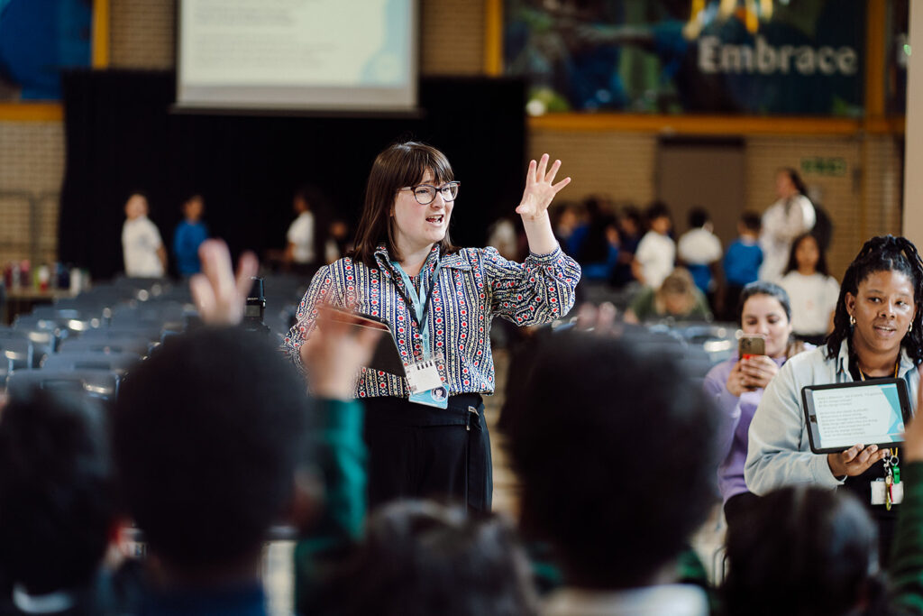 A teacher with a lanyard gestures enthusiastically during a classroom activity, engaging with a diverse group of students who are mostly out of focus in the foreground. Another educator assists by holding a tablet, while in the background, more students can be seen in a school hall with chairs and a presentation screen, creating an atmosphere of interactive learning.