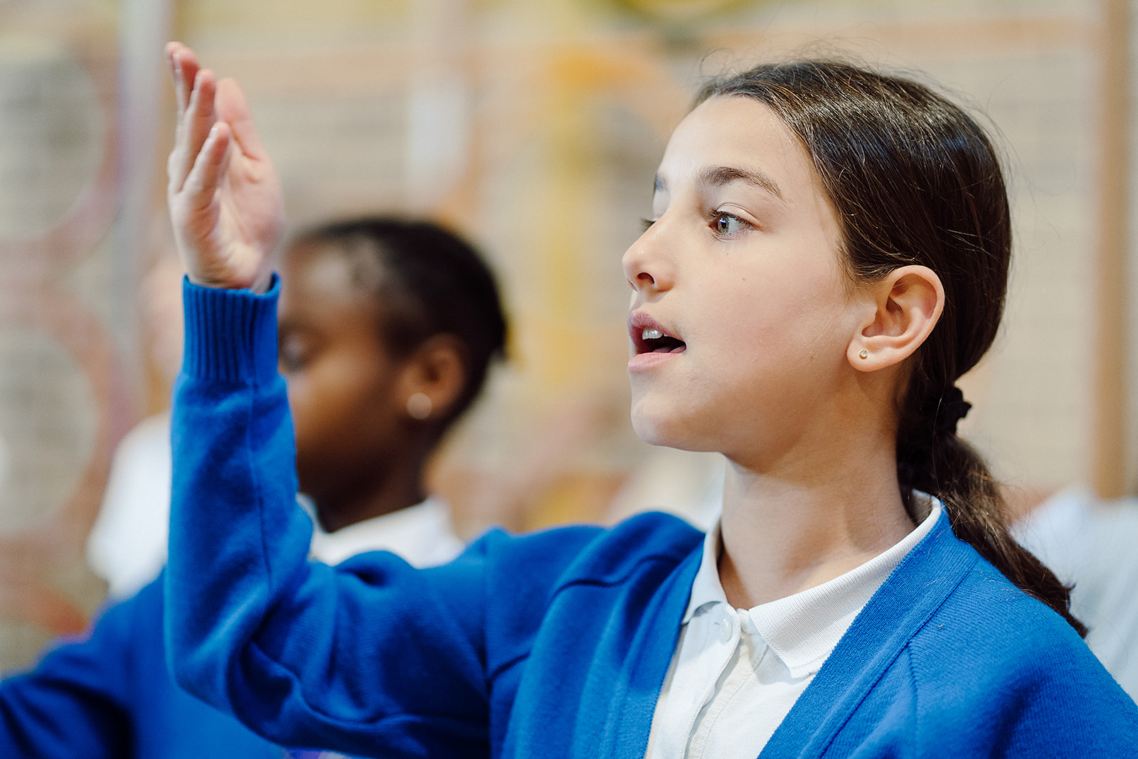 A young girl in a blue cardigan and white shirt is captured mid-song, her expression focused and joyful. Her raised hand suggests active participation in a choir or music class, while a blurred classmate in the background shares in the harmonious activity, illustrating the vibrant and collaborative atmosphere of a Sing Education musical learning experience.