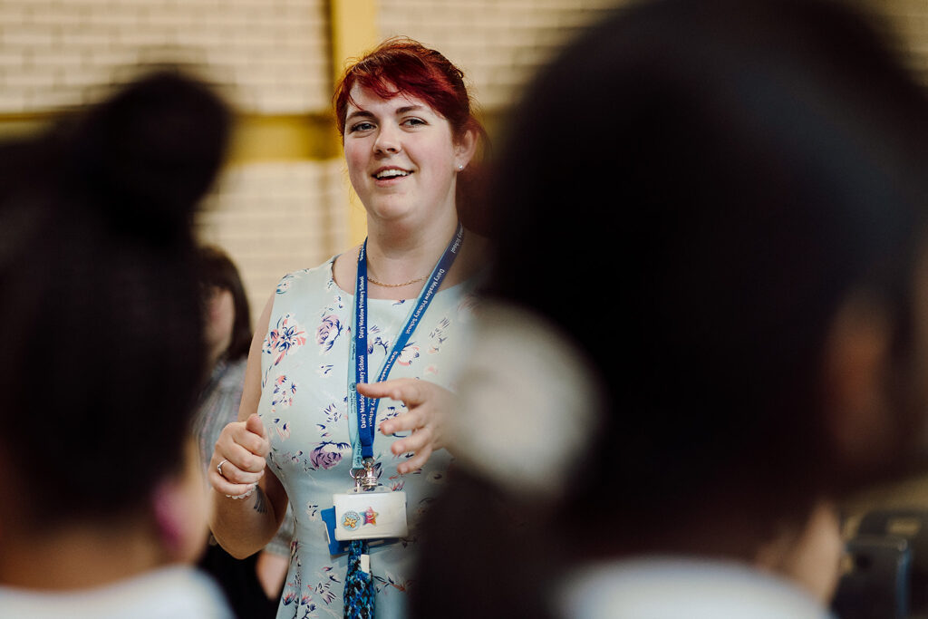 A smiling educator with red hair, wearing a floral dress and a lanyard, actively engages with students in a classroom setting, her hands expressively gesturing as she communicates with the out-of-focus students in the foreground, creating a sense of connection and dynamic interaction in the learning environment.