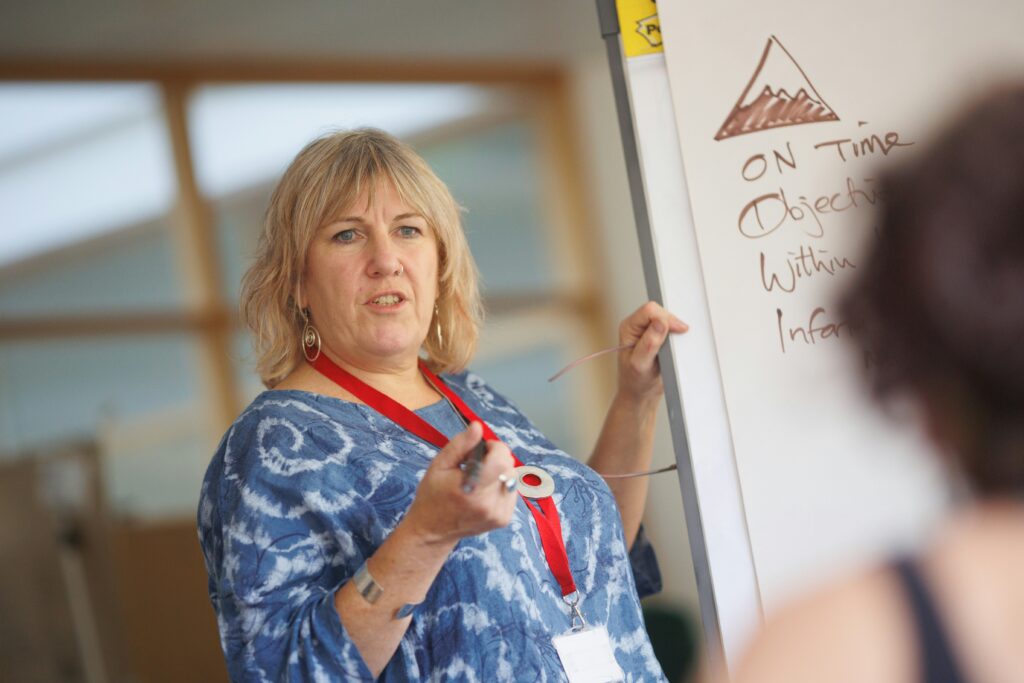 A woman in a blue top with a red lanyard is standing in front of a whiteboard, teaching a class.