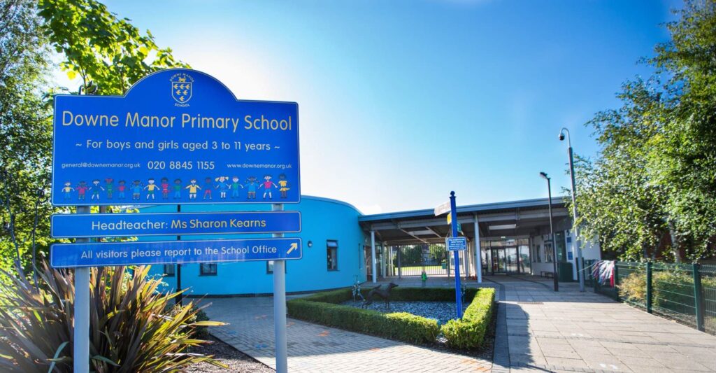 A primary school building painted blue is pictured on a sunny day. In the foreground a welcome sign shows this is "Downe Manor Primary School, for boys and girls aged 3 to 11 years" It also shows the headteacher's name is Sharon Kearns. The school looks welcoming and friendly with artwork and flags dotted around.