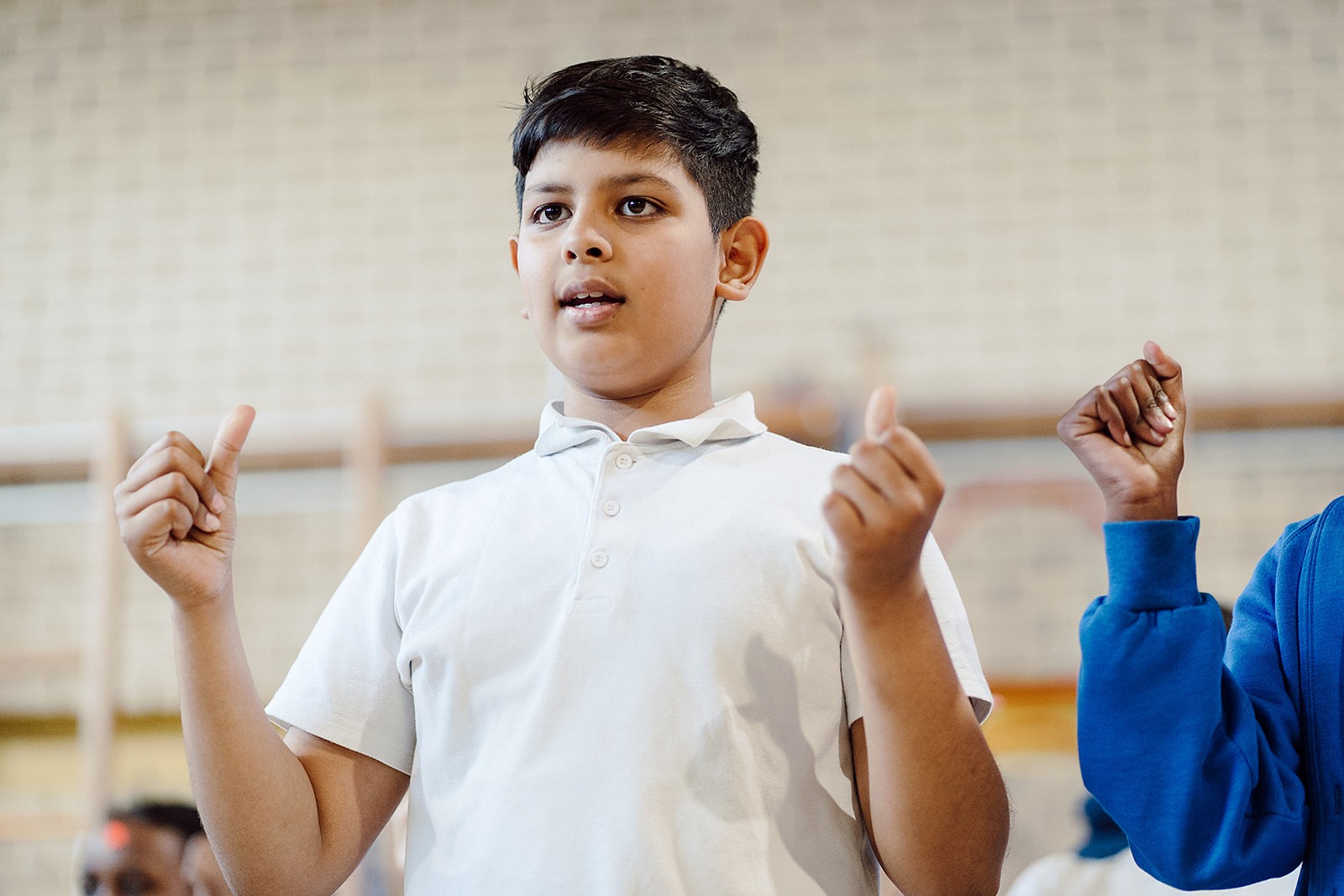 An older student clicking his fingers, is featured prominently in the foreground, wearing a school uniform with a white polo shirt. In the blurred background, classmates can be seen in the school hall, suggesting a lively singing assembly or performance. The image shows the positive impact that music is having on this student