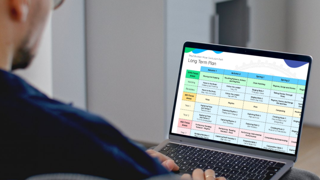 Looking over a man's shoulder at his computer. He is looking at the Sing Education long term plan which is part of a larger music curriculum pack