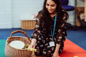 A smiling educator kneels on a colorful classroom rug, reaching into a woven basket filled with musical instruments, creating an inviting atmosphere for learning through music. Her engagement with the instruments suggests an interactive and joyful approach to teaching.