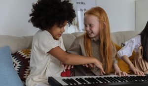 Two young children are sharing a joyful moment while playing together on a digital piano. The child with curly hair is smiling and pointing at the keys, while the red-haired child looks on with a bright, engaging smile, suggesting a playful and collaborative music-making experience. They are seated comfortably on a couch with colorful cushions, indicating a relaxed and friendly learning environment.